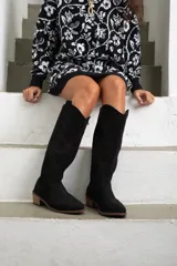 Bota de caña alta estilo western, confeccionada en gamuza negra con detalles bordados sutiles en la caña y el empeine. Cuenta con un taco bajo y suela de madera.