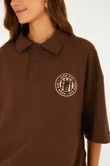 Remera tipo polo marrón, de corte oversize, con cuello clásico, cartera con botones y mangas amplias hasta el codo. Cuenta con un detalle bordado circular en el pecho.