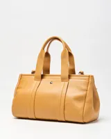 Cartera tipo tote XL confeccionada en símil cuero de textura granulada color habano. Cuenta con doble asa de mano, detalles de correas decorativas con hebilla en los laterales y un charm colgante en forma de corazón. Incluye placa metálica con las iniciales de la marca en el frente.