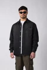 Chaqueta de denim negro con efecto desgastado, corte relajado, cierre frontal con botones metálicos, cuello clásico y bolsillo frontal en el pecho.