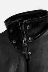 Cazadora de piel 100% color negro de Zara Woman Collection, con cuello subido, manga larga y bolsillos de plastrón con solapa en el delantero. Cuenta con bajo elástico y cierre frontal con cremallera y botones a presión ocultos.
