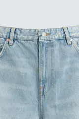 Bermudas de denim de algodón, color celeste claro con efecto lavado y manchas marrones/beige en las perneras. Tienen un corte ancho (baggy fit) y terminación de bajo irregular.