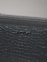Tarjetero Calvin Klein color gris oscuro con textura de piel de cocodrilo sintética grabada. Cuenta con cuatro ranuras para tarjetas, dos bolsillos con ranura y un bolsillo monedero con fuelle y cremallera. El logo de Calvin Klein es de metal plateado brillante en la parte delantera.