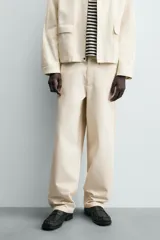 Pantalón chino de algodón color beige, de corte relajado (relaxed fit) y tiro medio. Presenta bolsillos delanteros y bolsillos traseros de vivo con cierre de cremallera y botón.