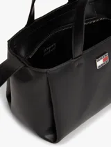 Cartera tote de poliuretano sintético liso color negro de la marca Tommy Jeans, con dos asas superiores y correa ajustable. Presenta cierre de cremallera, compartimento principal e insignia metálica de la marca en la parte delantera. Incluye un adorno colgante extraíble con cordón azul trenzado, dije en forma de corazón y herrajes plateados.
