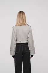 Trinchera corta color gris claro, con solapas, doble botonadura, cinturón y mangas largas con trabillas ajustables.