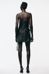 Vestido corto de escote fluido y tirantes finos, completamente cubierto de lentejuelas negras. Presenta detalles de volantes en tejido combinado que recorren el lateral y el frente de la prenda.