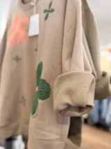 Buzo de cuello redondo en color beige, con diseño oversize y felpa interna. Presenta bordados de flores en tonos rosa, verde y azul distribuidos en el frente.