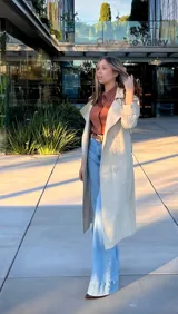 Trench coat de color beige, con diseño de doble botonadura, solapas amplias y cinturón ajustable en la cintura. Cuenta con mangas largas con trabillas en los puños y bolsillos laterales.