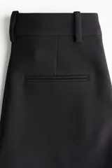 Pantalón negro de tiro alto con pierna ancha y pliegues marcados al frente. Presenta cierre frontal y un botón decorativo dorado en la pretina.