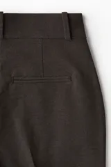 Pantalón sastrero de tiro alto con cierre frontal, botón oculto y corchetes. Presenta bolsillos al bies, bolsillos decorativos traseros y piernas de corte recto con pliegues marcados tanto en el frente como en la parte posterior.