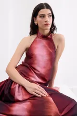 Vestido largo de corte holgado con estampado degradé en tonos rojizos y bordó. Presenta escote halter con cuello alto, espalda descubierta con cierre de botones y un maxi volado en la cadera que aporta volumen a la falda.