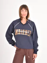 Buzo azul oscuro de corte holgado con cuello polo y escote en V. Presenta mangas largas con costuras raglán en color blanco y un estampado frontal con la palabra Ambience y el texto United Nation League en tonos dorados.