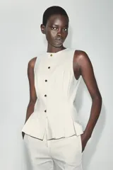 Chaleco entallado blanco con cuello redondo, manga sisa, pinzas estructuradas, detalle de peplum en la parte inferior y cierre frontal con botones dorados.