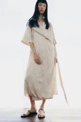 Vestido midi de Zara Woman Collection color beige, con cuello solapa, mangas por debajo del codo y diseño cruzado. Confeccionado en tejido plisado con brillo satinado, incluye un cinturón ajustable del mismo género con lazada en la cintura.