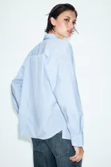 Camisa de manga larga con patrón de rayas verticales en tonos celeste y blanco, cuello con solapa abotonada y un bolsillo aplicado en el pecho. Cierre frontal con botones blancos.