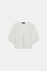 Chaqueta corta de punto color blanco crudo, con escote redondo pronunciado y cierre frontal con botones oscuros. Presenta mangas cortas abullonadas con frunces.