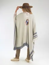 Poncho de hilo color crudo, con diseño de rayas azules y flecos en los bordes. Cuenta con un escudo bordado en la parte posterior.