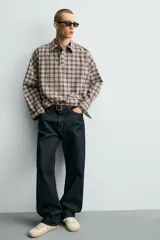 Camisa polo de corte boxy fit confeccionada en tejido de algodón con elasticidad, presenta un estampado de cuadros en tonos marrones, azules y blancos. Cuenta con cuello solapa, cierre frontal de botones, manga larga con puños abotonados y bajo con aberturas laterales.