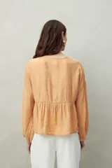 Blusa de manga larga color beige claro, con escote en V y lazos ajustables en el centro del pecho. Presenta frunces horizontales que generan volumen en el busto y el torso.