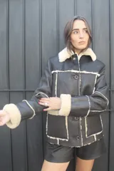 Chaqueta tipo gamulán de cuero genuino en color negro, con interior y detalles de corderito en color crema. Presenta cierre frontal con botones, cuello solapa y diseño de paneles con costuras a la vista.