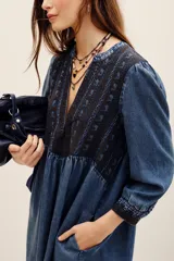 Vestido corto de tencel en color azul, con mangas largas abullonadas y escote en V profundo. Cuenta con detalles de bordados oscuros en el pecho, los puños y el ruedo, y una falda de corte evasé.