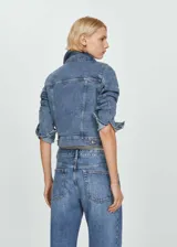 Chaqueta de jean estilo vaquero, con diseño recto y corto. Presenta cuello de solapa, cierre frontal de botones metálicos, dos bolsillos con solapa en el pecho y dos bolsillos laterales. Las mangas son largas con puños abotonados.