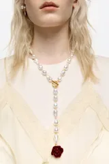 Collar largo tipo lazo compuesto por perlas irregulares de vidrio, con cierre frontal de barra y argolla en tono dorado. Finaliza en una sección colgante que termina con un dije en forma de rosa roja de resina.