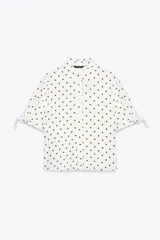 Camisa de manga corta confeccionada en tejido de ramio, color blanco con estampado de lunares marrones. Presenta cuello solapa, cierre frontal con botones y detalle de lazos en el acabado de las mangas.