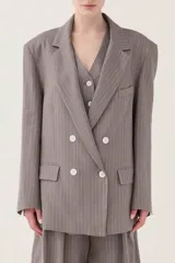 Blazer oversize cruzado color gris con rayas diplomáticas blancas, hombros marcados, solapas anchas y cierre de botones blancos. Cuenta con bolsillos laterales con solapa y un bolsillo ojal en el pecho.
