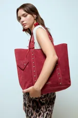 Cartera tipo shopper de gran tamaño confeccionada en material efecto gamuza color rojo. Presenta correas reforzadas decoradas con tachas metálicas plateadas y un dije colgante en forma de corazón con detalles de tachas. Cuenta con una placa metálica pequeña con logo en la parte inferior frontal.