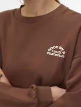 Buzo boxy marrón con cuello redondo, manga larga y diseño holgado, con pequeño estampado circular en el pecho.