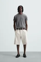 Bermuda color beige claro, de corte ancho y largo, confeccionada en algodón. Presenta cierre frontal con cremallera y botón, bolsillos laterales, bolsillo cerillero frontal y bolsillos traseros de vivo.