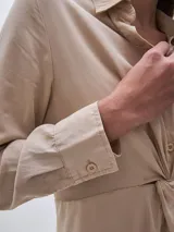 Vestido camisero largo color beige, con cuello clásico, mangas largas y cierre frontal con botones. Presenta un nudo decorativo a la altura de la cintura y una abertura en el ruedo.