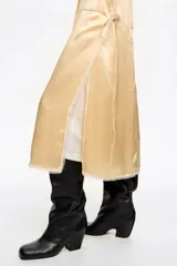 Vestido midi de diseño bimateria con sobrevestido de satén color amarillo claro. Presenta escote en V profundo con lazo frontal, detalles de encaje en el ruedo y aberturas laterales con cordones. Se combina con una base de remera blanca de manga corta y cuello redondo.