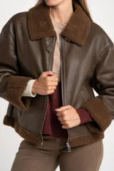 Campera tipo gamulán de cuero sintético en color marrón, con interior y detalles en cuello y puños de corderito sintético al tono. Presenta un corte oversize con cierre frontal metálico.