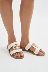 Sandalias planas color marfil con dos tiras anchas en el empeine, decoradas con un aplique metálico dorado de diseño orgánico en el centro. Suela marrón.