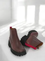 Bota corta de cuero marrón con suela track negra, cierre lateral y detalle de tira de tela roja en la parte trasera.