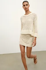 Minifalda de tiro medio confeccionada en ante color beige, decorada con aplicaciones de tejido tipo crochet blanco formando un patrón de rombos. Cuenta con cierre de cremallera oculta en la costura.