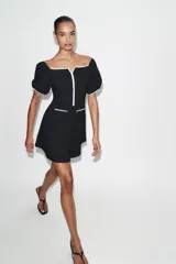 Vestido corto de tejido bouclé en color negro, con escote recto y mangas cortas abullonadas. Presenta detalles de vivos en color blanco a contraste en el escote, el centro del delantero y en los falsos bolsillos frontales.