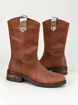 Bota de caña alta de cuero marrón, con taco bajo y detalles plateados en relieve texturizado. Cuenta con una hebilla metálica grande y tiras desmontables con tachas decorativas alrededor del tobillo, además de una presilla superior en la parte trasera.