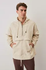 Campera canguro de gabardina color beige, con capucha ajustable con cordones, cierre frontal medio y bolsillo tipo ojal con solapa en el frente. Presenta mangas largas con puños elásticos y diseño holgado.