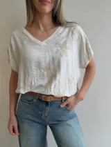 Blusa blanca de corte relajado, con escote en V, mangas cortas amplias y diseño oversize. Presenta delicados bordados florales en relieve distribuidos por toda la prenda.