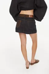 Minifalda de denim negro con costuras contrastantes en color ocre, pretina alta con trabillas y doble botón metálico.