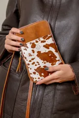 Pocket bag de cuero vacuno con diseño combinado en gamuza color tostado y cuero con pelo estampado animal print tipo vaca. Cuenta con bolsillo frontal con detalle de bordado metálico, cierre superior y correa desmontable.