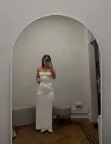 Vestido largo blanco elastizado, con escote recto y tirantes finos. Presenta una sobrefalda asimétrica con frunce lateral y abertura, y un estilo corset en la espalda.