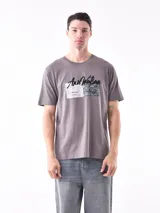 Remera de manga corta color gris topo, corte slim, con cuello redondo y un estampado gráfico en el pecho que combina texto manuscrito con un diseño abstracto en tonos oscuros.