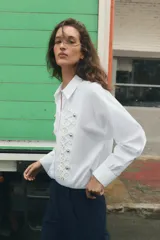 Camisa de popelín blanca con cuello solapa y manga larga. Presenta un diseño frontal con bordados en relieve y botones decorativos negros. Cierre frontal con botones ocultos.