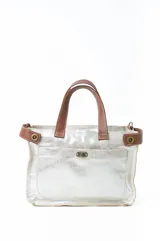 Bolso de mano tipo tote, confeccionado en cuero metalizado color plateado con detalles en cuero marrón en las asas y laterales. Cuenta con un bolsillo frontal con cierre giratorio y herrajes metálicos.