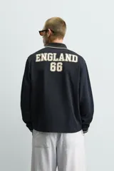 Polo de manga larga en tejido técnico acanalado color negro, con cuello solapa y abertura frontal. Presenta puños con ribete a contraste y una etiqueta aplicada en el pecho con el diseño conmemorativo de la Copa Mundial de la FIFA 1966.